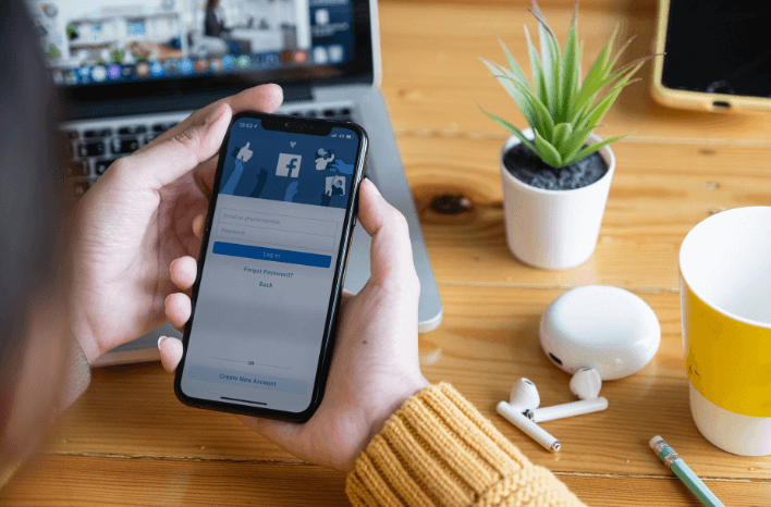 smartphone held in person's hands. phone is open to facebook. laptop computer, plant, and headphones visible on the desk.