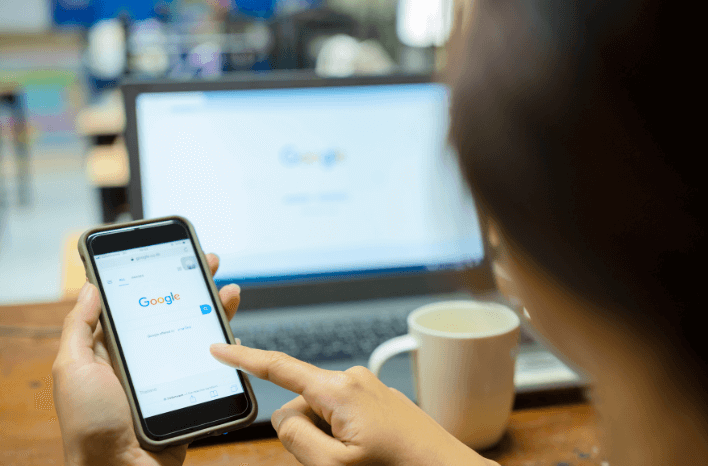 Person starting a Google search on their phone with the computer open to Google.com in the background | Truth Tree knows school marketing | Truth Tree blog post about school SEO in response to Google's leaked search ranking data | Truth Tree is a digital marketing agency working exclusively with schools