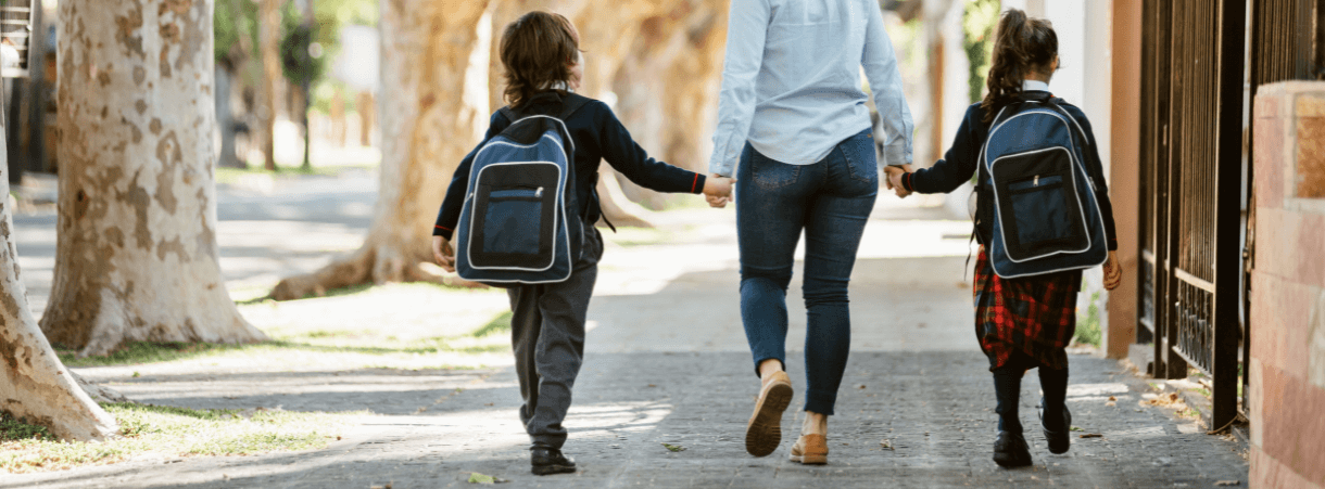 a woman wearing jeans and a top walks down the street holding the hands of a boy in uniform and backpack on the left and the hand of a girl in uniform and backpack on the right | image accompanies a Truth Tree page distilling survey data on newly enrolled families