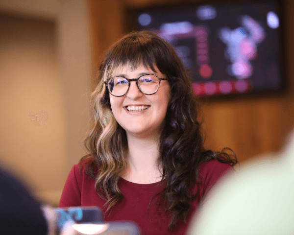 woman wearing glasses smiles at the camera with digital screen on the wall behind her