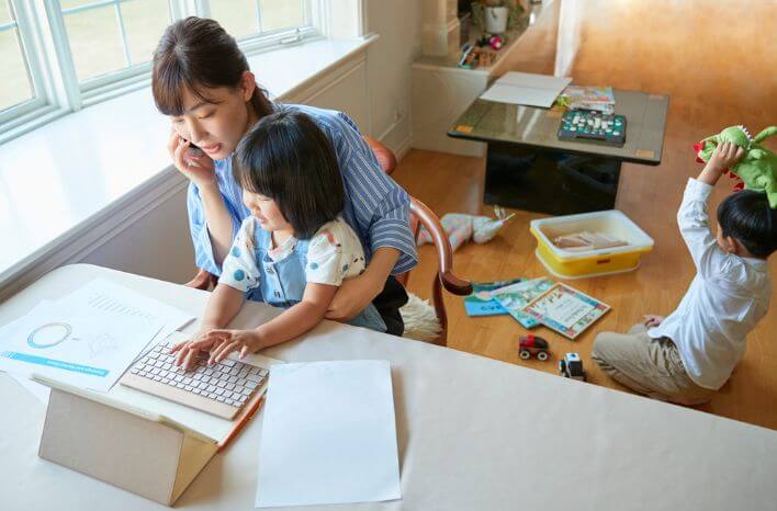 Busy mother on a phone call at her laptop with a toddler on her lap and another child playing nearby. SEO for schools comes down to one question, shown in the text on the image: “She’s juggling two kids and a job. When she finally has five minutes to research schools, will she find yours?”